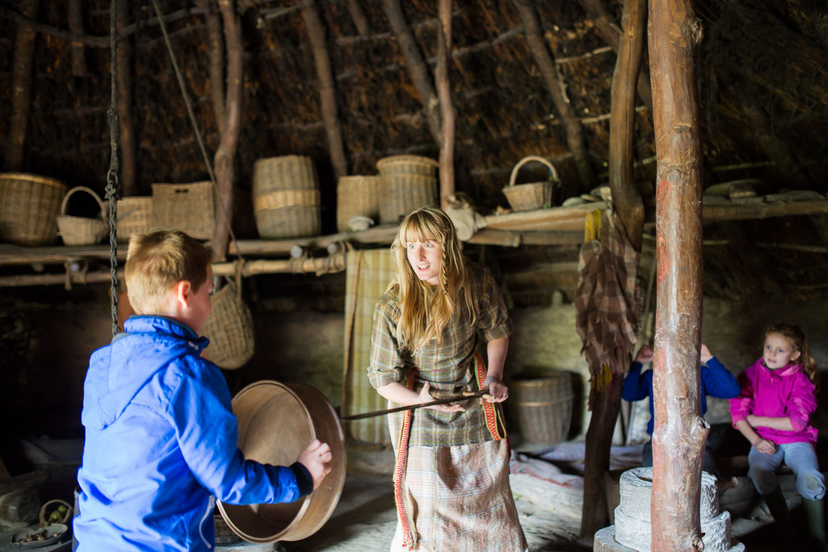 A costumed guide inside a dim roundhouse at Castell Henllys Iron Age Village is speaking to a young boy with baskets on shelf in the background