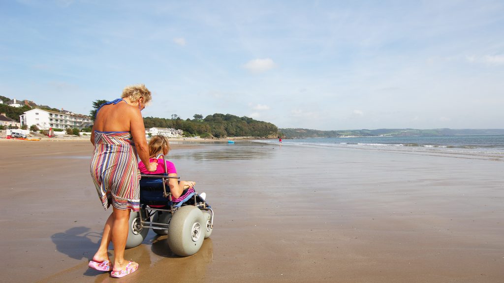 Adult woman pushing young woman in beach wheelchair along sandy beach on a sunny day