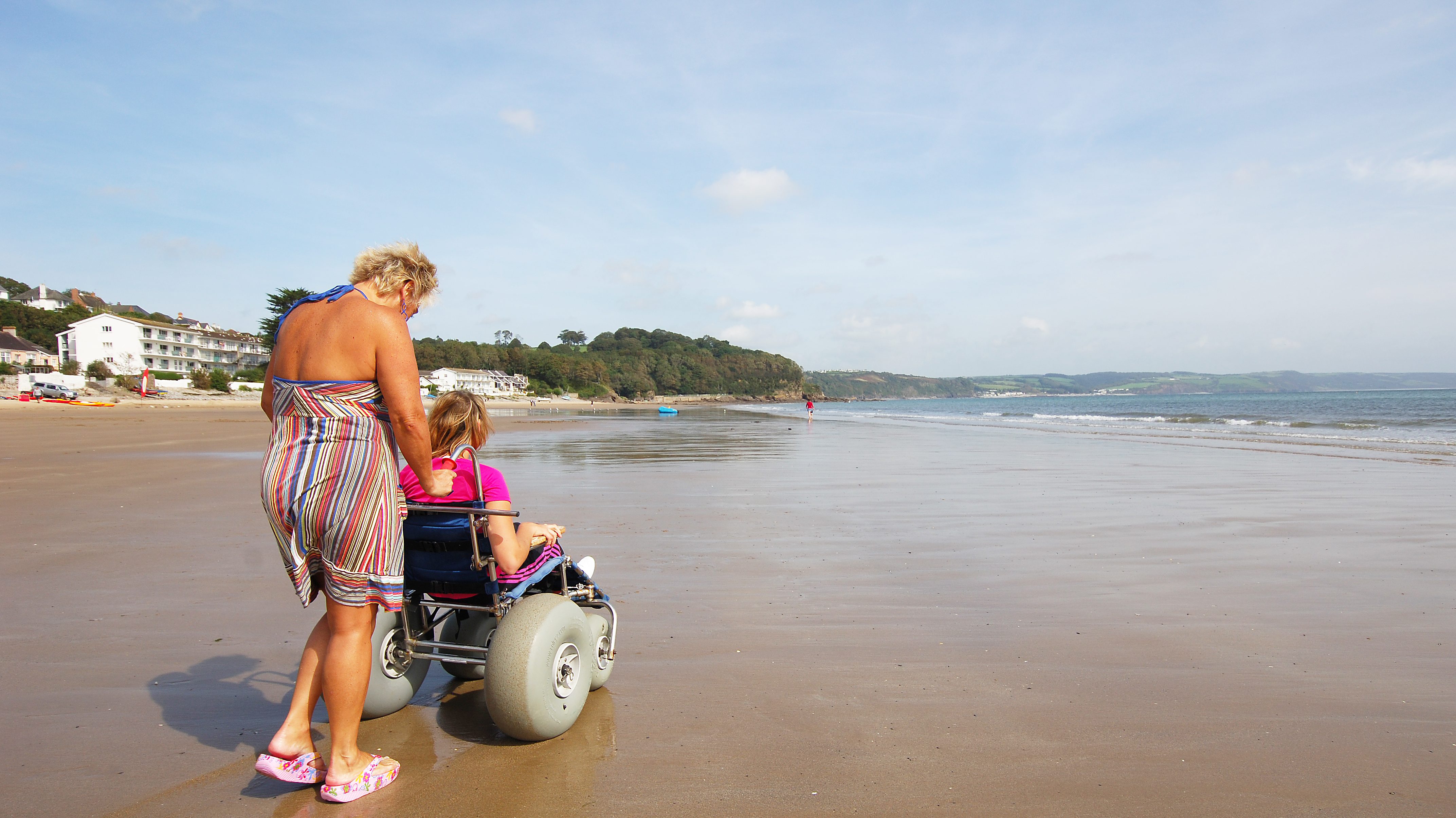 Adult woman pushing young woman in beach wheelchair along sandy beach on a sunny day