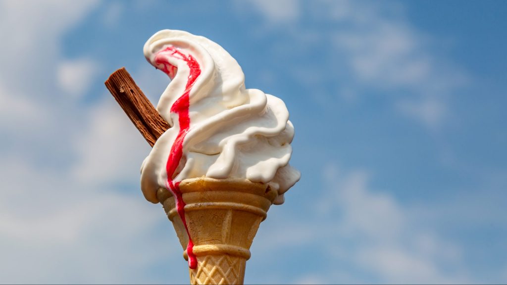 A vanilla ice cream cone with strawberry sauce, against a blue sky