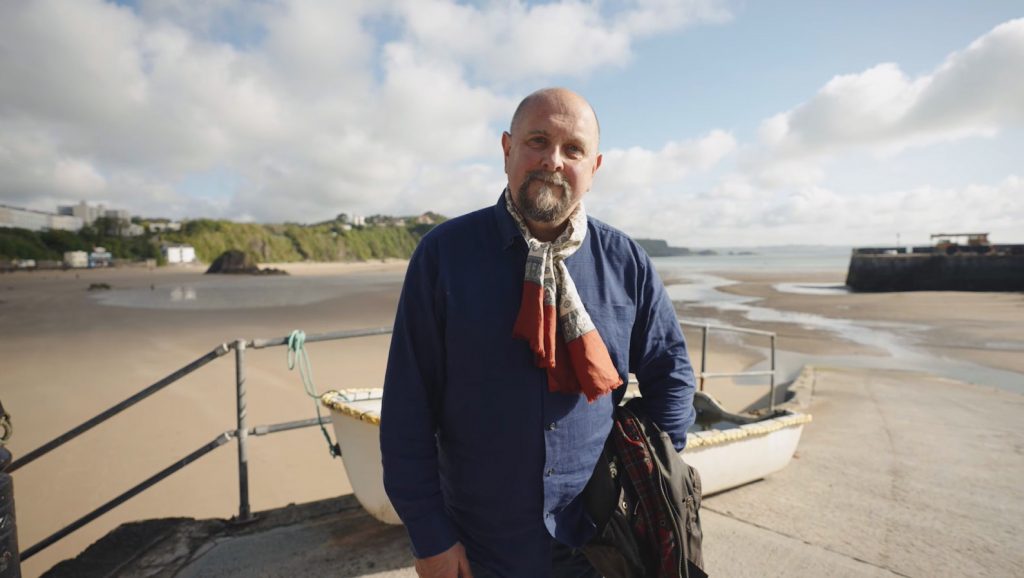 middle aged white man with goatee beard standing on a harbour wall above a sandy beach