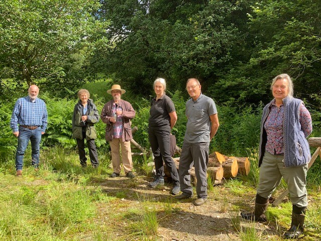 6 people standing in a wooded area next to a pile of sawn logs