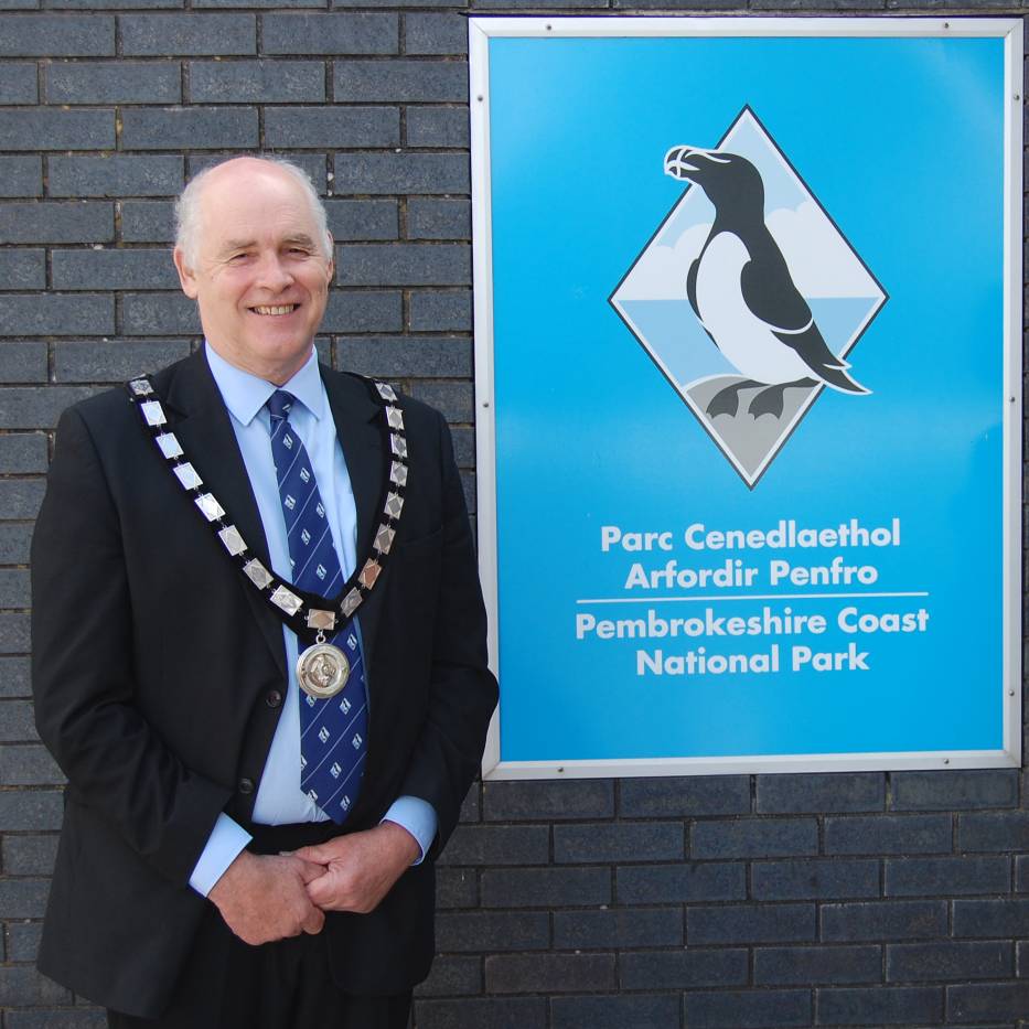 Man in suit wearing ceremonial tie standing alongside a sign that reads 'Pembrokeshire Coast National Park'