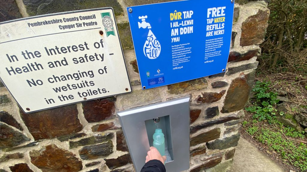 Arm holding an empty water bottle to an exterior refill tap