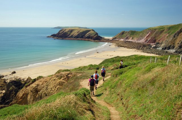 Group of people walking on a coastal footpath in single file with a sandy beach and rocky cliffs in the background