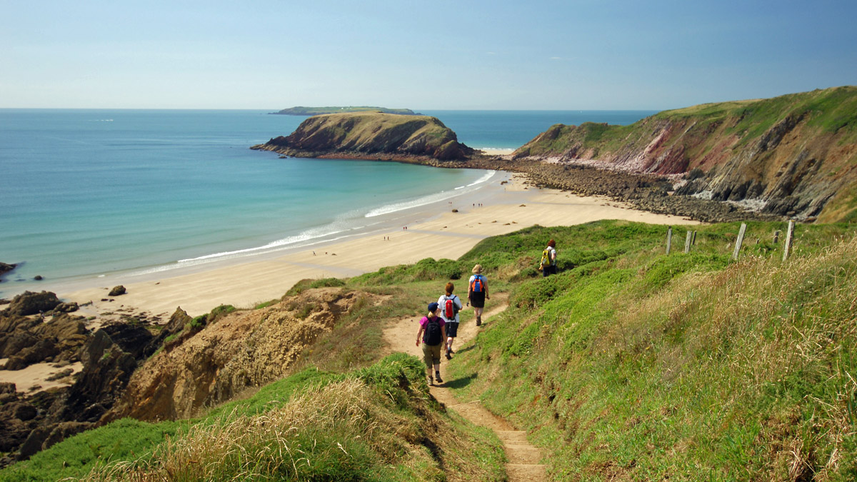 Group of people walking on a coastal footpath in single file with a sandy beach and rocky cliffs in the background