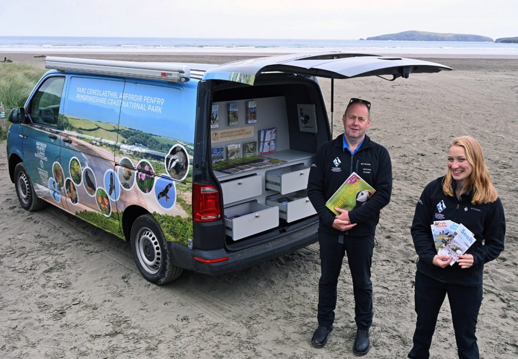 Two people holding information leaflets standing next to a converted VW van parked at the entrance to a sandy beach. People pictured are Pembrokeshire Coast National Park Authority Rangers, location featured is Poppit Sands, Pembrokeshire