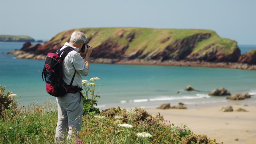 Man standing on grassy cliff edge taking photograph with camera. Location pictured is Marloes, Pembrokeshire, Wales, UK.
