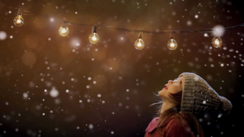 Girl wearing a winter hat looking up towards a series of string lights