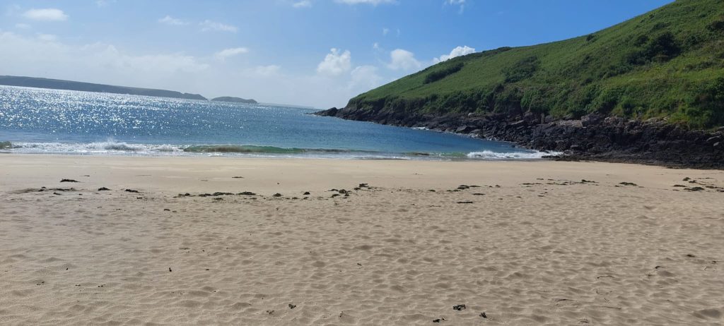 Sandy beach on a sunny day with grassy headland jutting out into the sea to the right hand side. Location is Watwick Bay, Pembrokeshire
