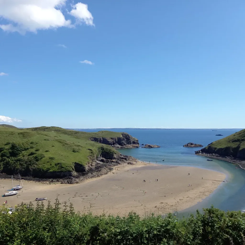 Sunny day photograph of an opening to a sandy harbour flanked by grassy cliffs either side. Location is Solva, Pembrokeshire.