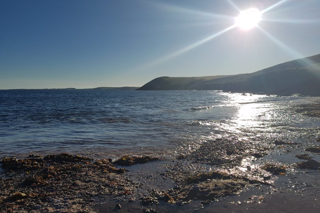 View across a beach on a sunny day with a view across the shallows to a headland jutting out into the sea in the distance. Location pictured is Manorbier, Pembrokeshire