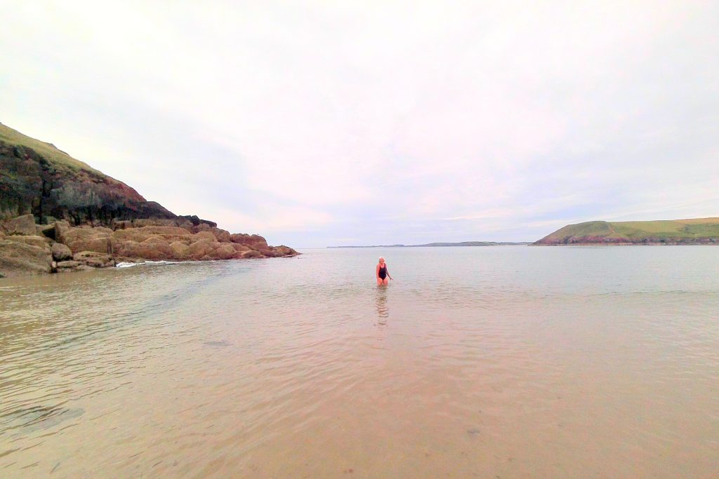 Single female in a one piece bathing suit standing in knee-high water at a sandy beach. Sandstone cliffs can be seen to the left and right. Location pictured is Manorbier, Pembrokeshire