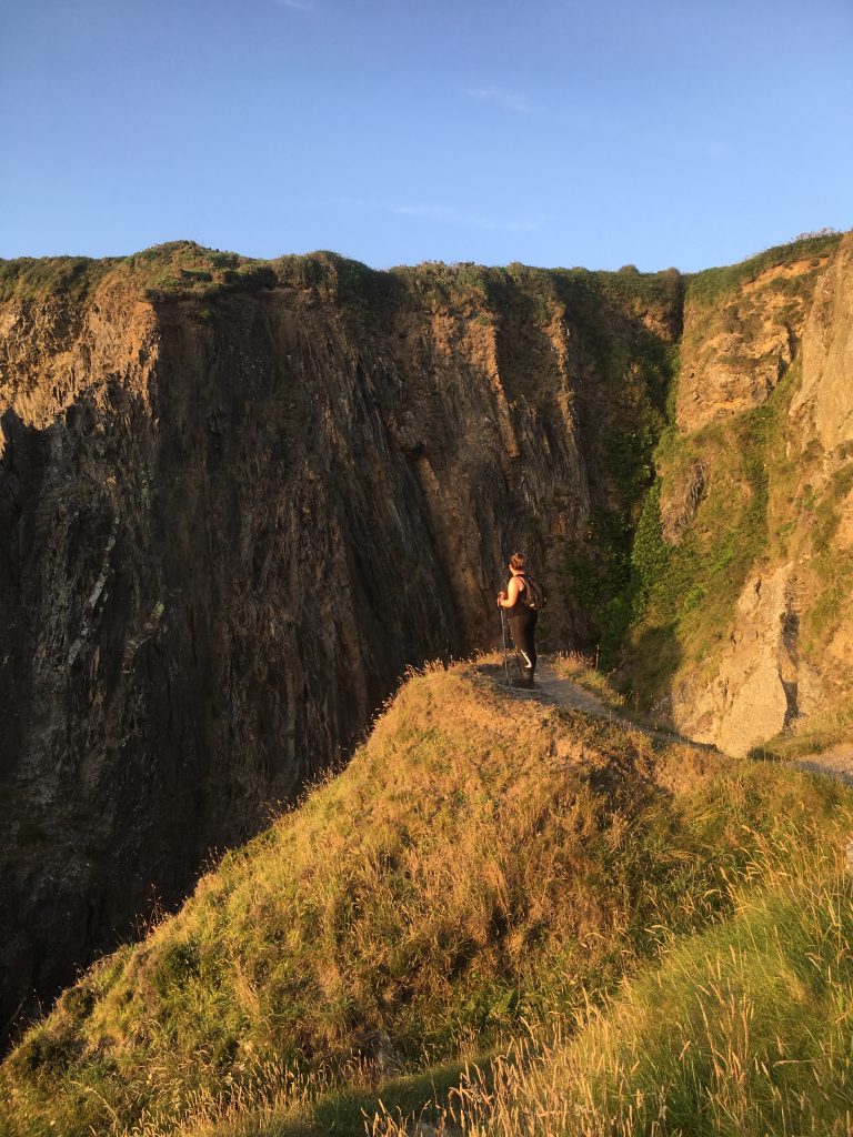 Single person standing on the edge of a grassy headline looking out across a void to rocky cliffs opposite. Location is between Pwll Deri and Aberbach on the Pembrokeshire Coast Path