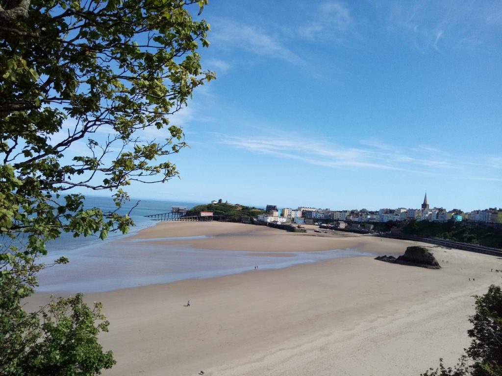View across a sandy beach to a seaside town featuring a harbour, two lifeboat houses, colourful townhouses anda church spire towering above them. Location pictured is Tenby, Pembrokeshire