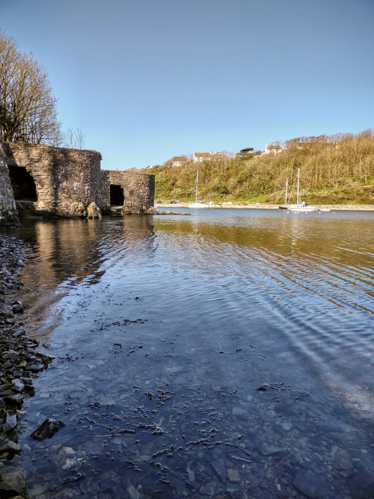 Two lime kilns located on the left hand side of a harbour with boats moored in the distance and a wooded cliff behind with houses perched atop the cliff. Photograph is taken across the shallow water at low tide. Location pictured is Solva, Pembrokeshire