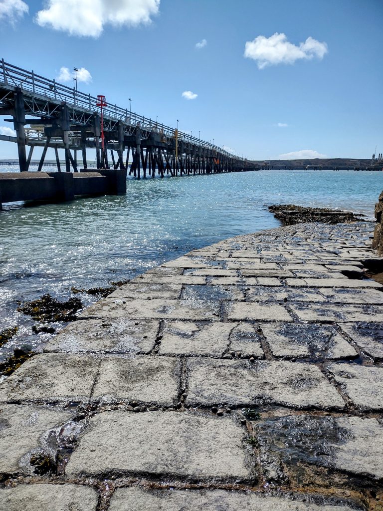 View from beneath a pier structure jutting out into a body of water with pipes running along one side of the structure. location is South Hook, Pembrokeshire