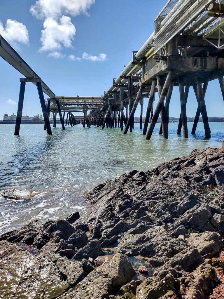 View from beneath a pier structure jutting out into a body of water with pipes running along one side of the structure. location is South Hook, Pembrokeshire