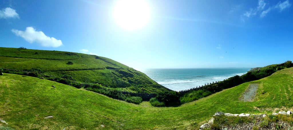 View of a beach from a grassy field behind a stone wall on a sunny day with hardly a cloud in the sky.  Location pictured is Druidston, Pembrokeshire