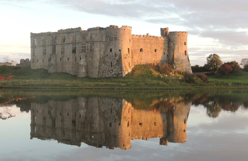 Partially ruined stone castle reflecting in the millpond it overlooks on an overcast day with the orange of a setting sun reflecting on one side of the castle walls. Location featured is Carew Castle, Pembrokeshire