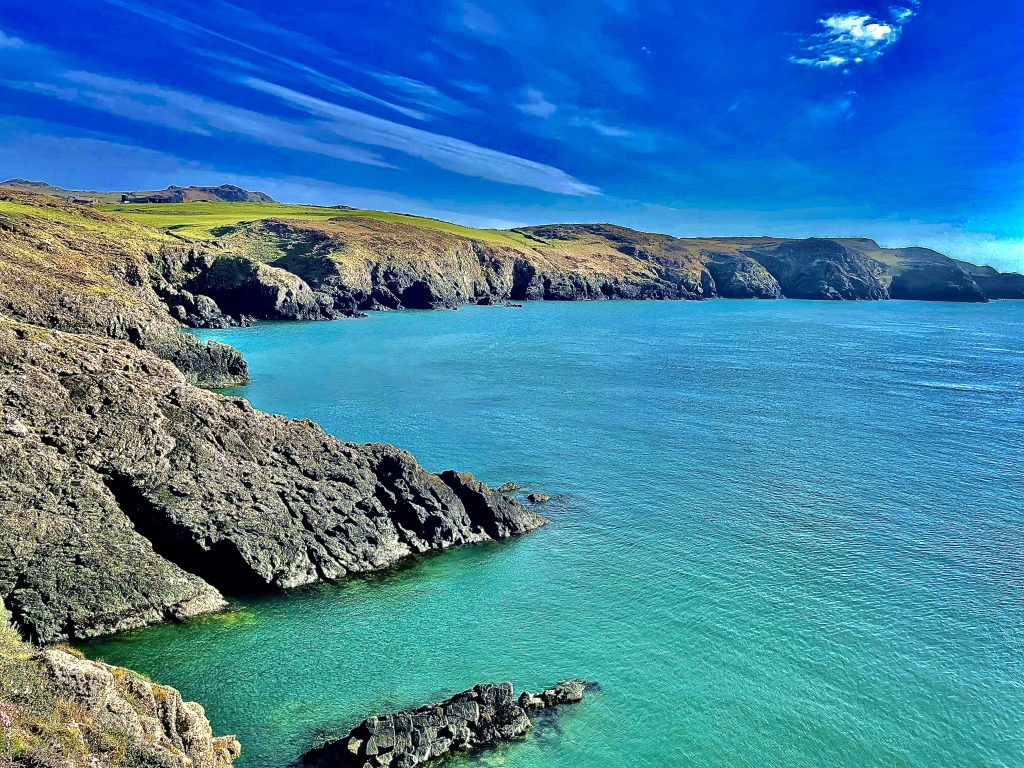 High contrast image of a rugged rocky coastline topped with grassy fields above aquamarine coloured seas on a sunny day.