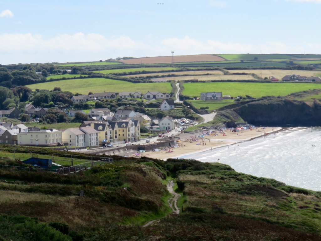 View of a small seaside town next to a sandy beach surrounded by grassy fields. The beach is busy with people enjoying a sunny day