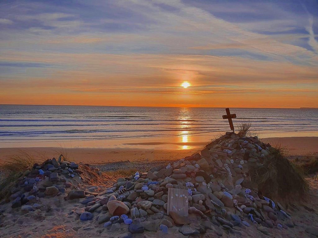 The sun setting above a sandy beach creating an orange glow across the sky and sea. In the foreground a pile of pebbles has been added to small sand dune mound with a small crucifix as its top. This is to mark the apparent site of Dobby's Grave, a fictional character from the Harry Potter books.