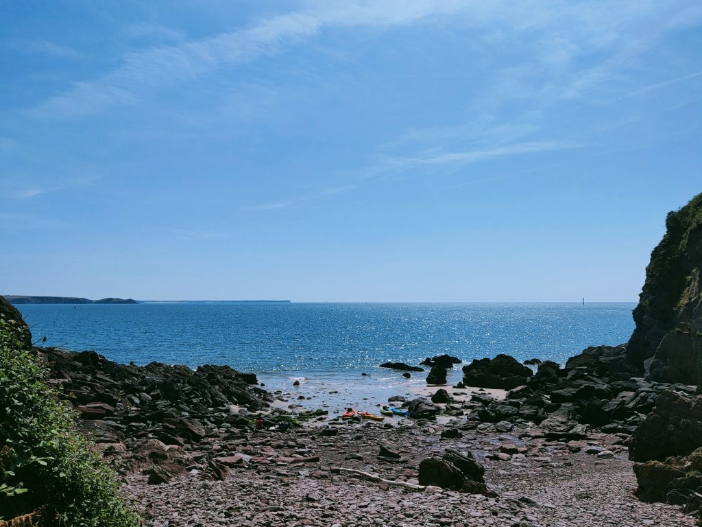 View from a rocky beach out to see, with four kayaks lying on the sandy area of the beach where the sea is lapping the shore. The sky is blue and sea is reflecting the sunny conditions. Location pictured is on the Dale Peninsula, Pembrokeshire