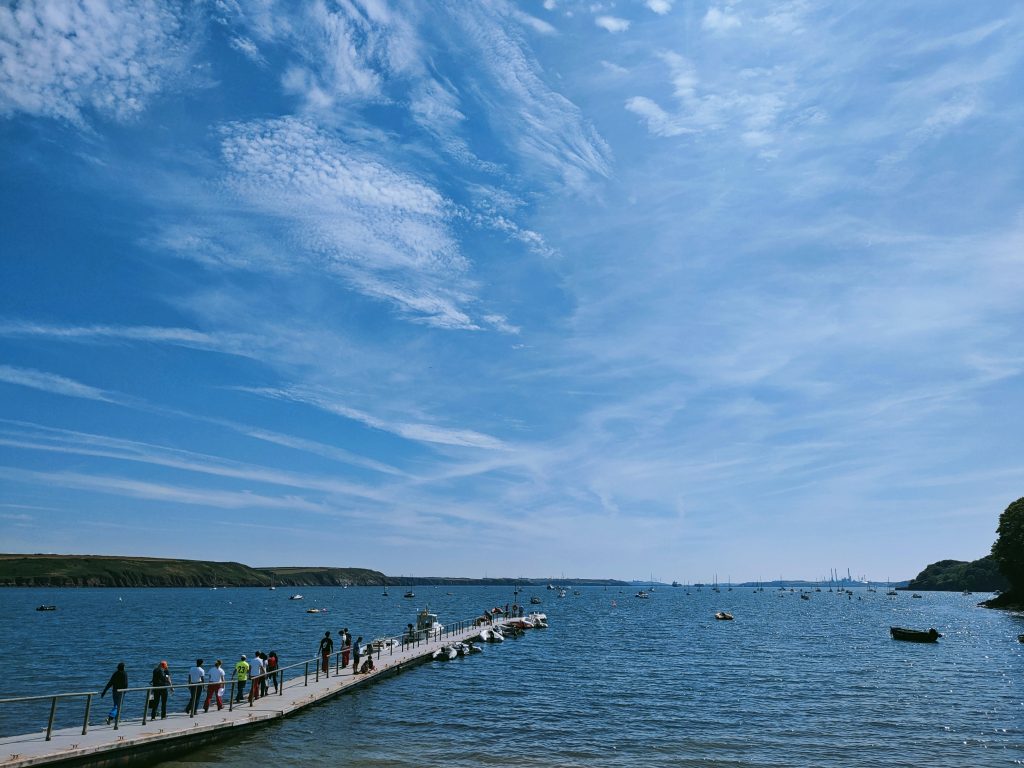 People walking along a floating pontoon where a number of small boats are moored and one larger boat awaits them. The pontoon is extending out into a relatively calm sea on a sunny day with blue skies and wispy clouds. In the distance you can see a number of boats moored out int the bay. Location pictured is Dale, Pembrokeshire