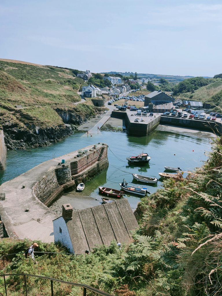 Photograph of a small fishing village with manmade harbour walls enclosing an area where several small boats are moored. Many cars are parked along the edge of the harbour wall and in the village behind it. Location pictured is Porthgain, Pembrokeshire