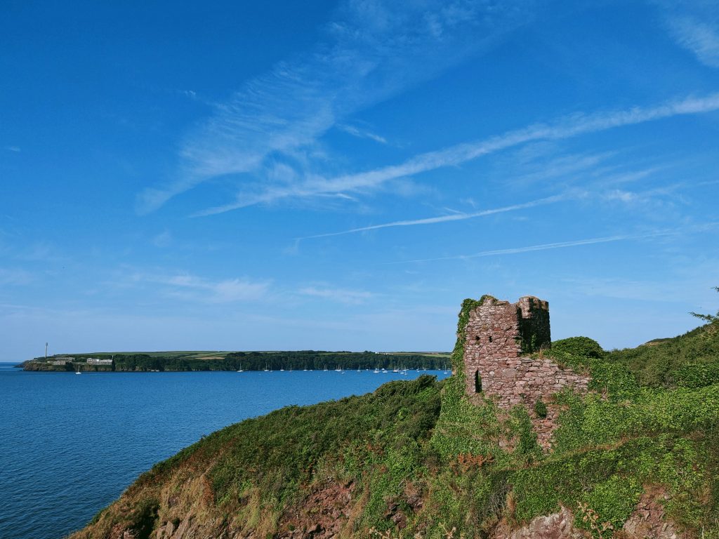 Ruined stone building on the edge of a cliff covered with grass and ivy with a view across to wooded coastline in the background with a number of boats moored next to the cliff. It's a sunny day with blue skies and blue seas. Location pictured is St Anne's Head, Pembrokeshire