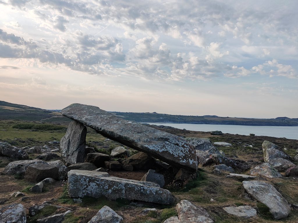 Remains of a stone burial chamber with one capstone being supported by two standing stones, one taller than another. There are a number of stones scattered around it and a view across the sea to a coastline in the background. Location pictured is Coetan Arthur on St David's Head, Pembrokeshire