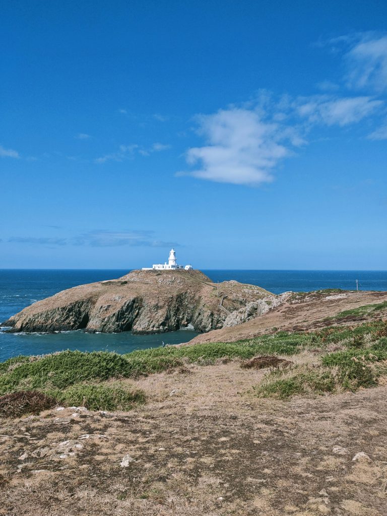 Lighthouse located on a rocky islet photographed from across a rugged clifftop on a sunny day with blue skies and blue seas below. Location pictured is Strumble Head, Pembrokeshire