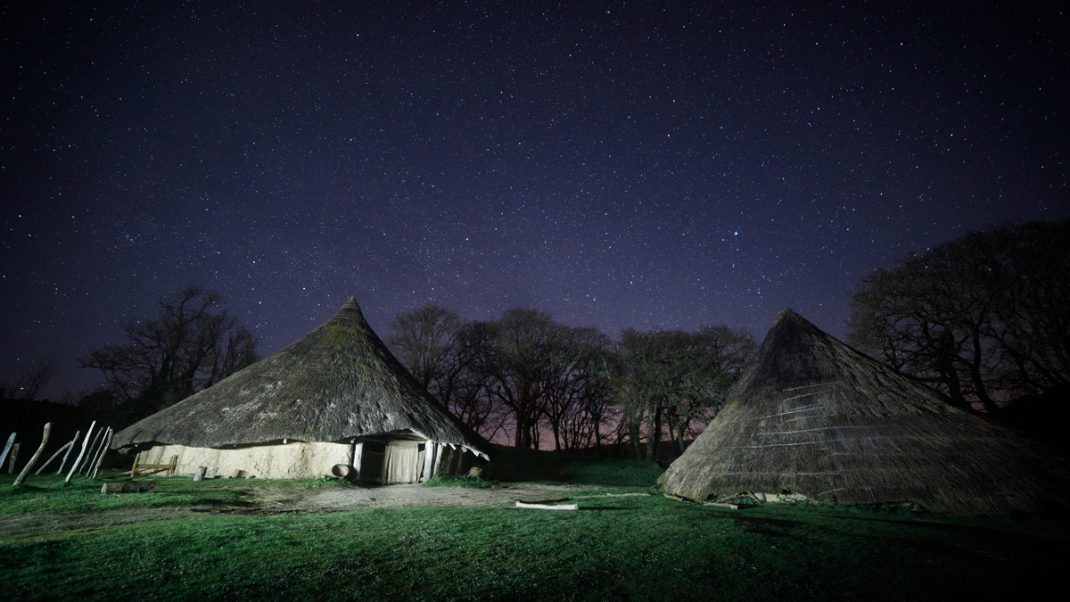 Reconstructed iron age roundhouse with a thatched roof pictured under a starry sky. Location pictured is Castell Henllys Iron Age Village, Pembrokeshire