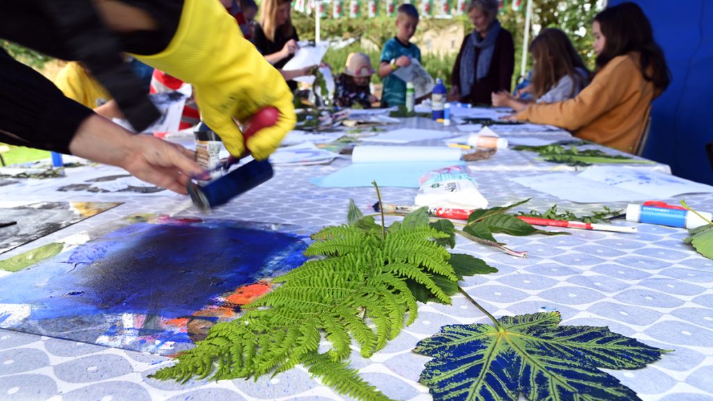 Range of art materials on table covered by a blue table cloth with people sat around creating collages.