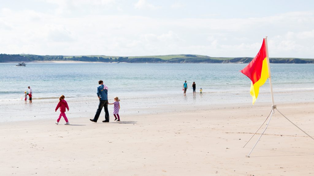 People walking along a sandy beach, passing by a red and yellow lifeguard flag. on a sunny day. Location pictured is Tenby, Pembrokeshire