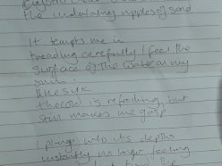 Image of a poem written on lined paper. It reads: Still seas. Light glistening on the surface like dappled gemstones. Crystal clear view down to the undulating ripples of sand. It tempts me in, treading carefully I feel the surface of the water on my skin Like silk, the cold is refreshing, but still makes me gasp  I plunge into its depths instantly no longer feeling the cold but find the warm sections on the surface caressed by the Sun.