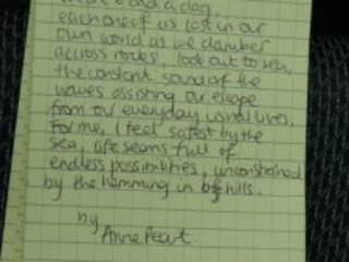 Poem written in a notebook. It reads: 4th August 2023. We are 6 and a dog Each one of us lost in our own world as we clamber across rocks, look out to sea,  The constant sound of the waves sound of the waves assisting our escape from our everday lives. For me, I feel safest by the sea, life seems full of endless possibilities, unconstrained by the hemming in of hills. By Anne Peart