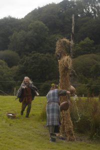 A woman in Iron Age costume pours beer at the foot of a large straw man