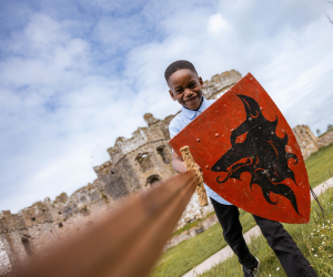 A young boy with a red shield outside Carew Castle, with his sword pointing towards the camera