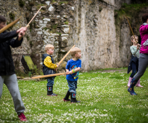 Children and an adult participate in a mock sword fighting lesson on a grassy field in front of a stone castle wall.