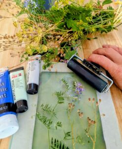 Overhead view of a table with a collection of wildflowers and greenery arranged around a printing plate, paint tubes (blue and light green), and a brayer.