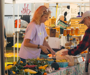 A vendor at an outdoor market hands a bag of produce to a customer, with a display of fresh vegetables, honey jars, and baked goods on the table.