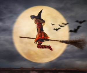 A child dressed as a witch on a broomstick flies in front of a large, glowing full moon against a cloudy night sky.