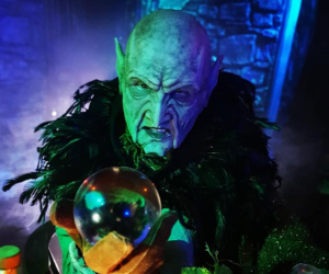 A scary, bald, green-skinned character with sharp teeth and dark feathers around its neck holds a crystal ball.