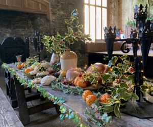 A long wooden table is laden with a festive spread of baked goods, roasted meat and garlands of holly and white lights, set against a stone wall and large windows.